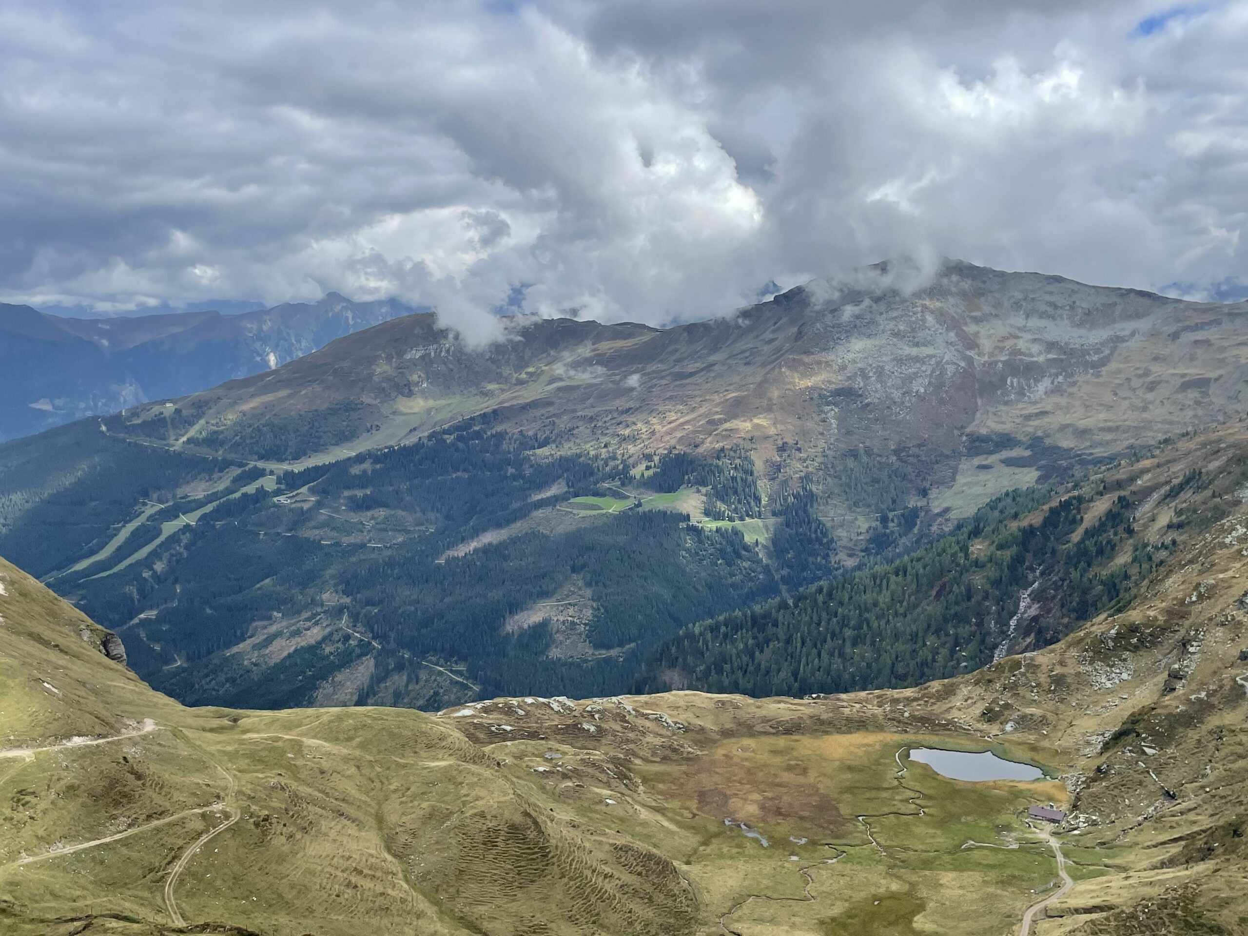 Weites Bergpanorama mit wolkenverhangenem Himmel, Waldhängen, Wegen und kleinem See im Tal. – Gadauner Alm