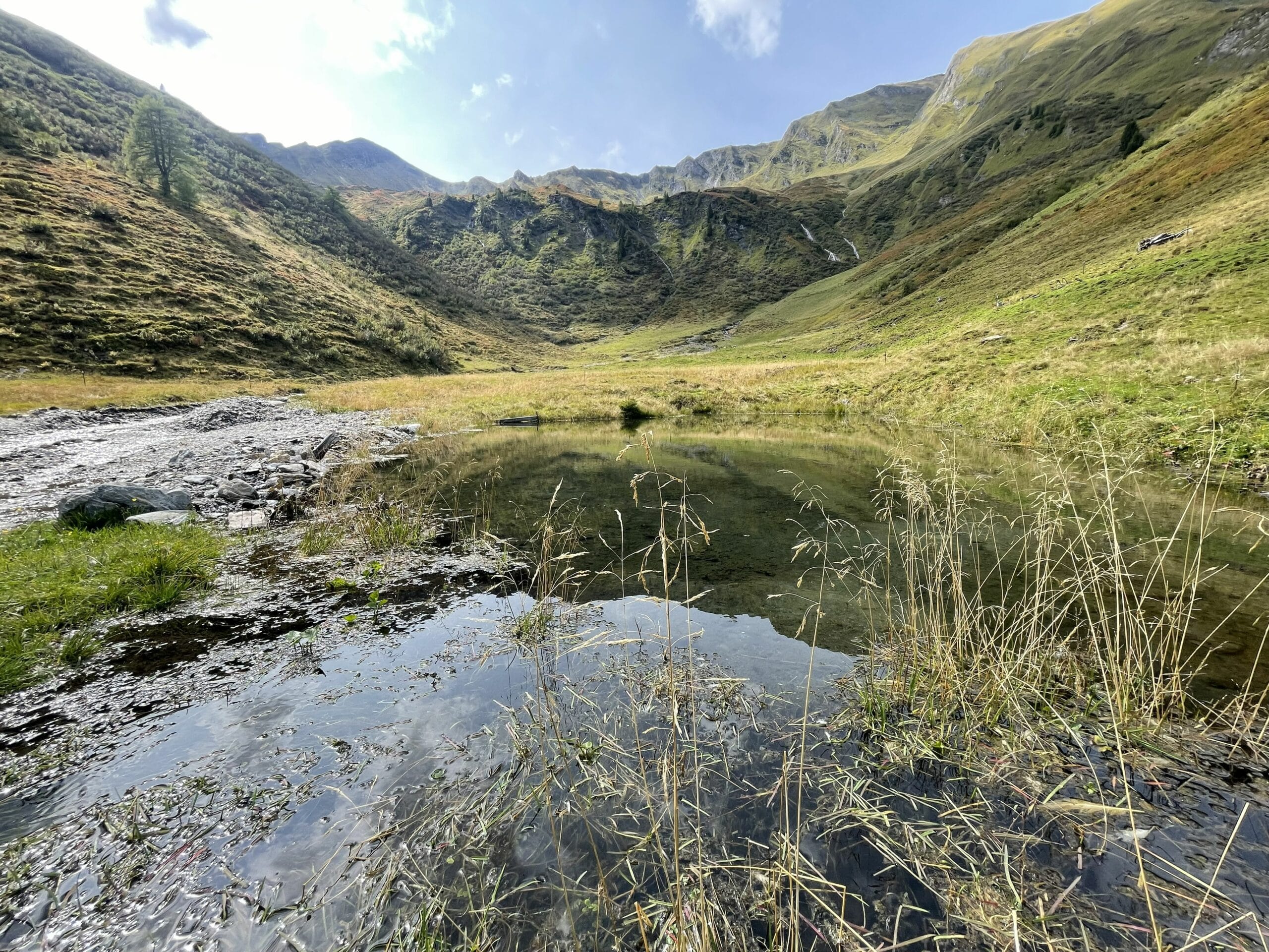 Bergtal mit kleinem Bach und ruhigem Wasserbecken, umgeben von grasigen Hängen und felsigen Bergen unter blauem Himmel.