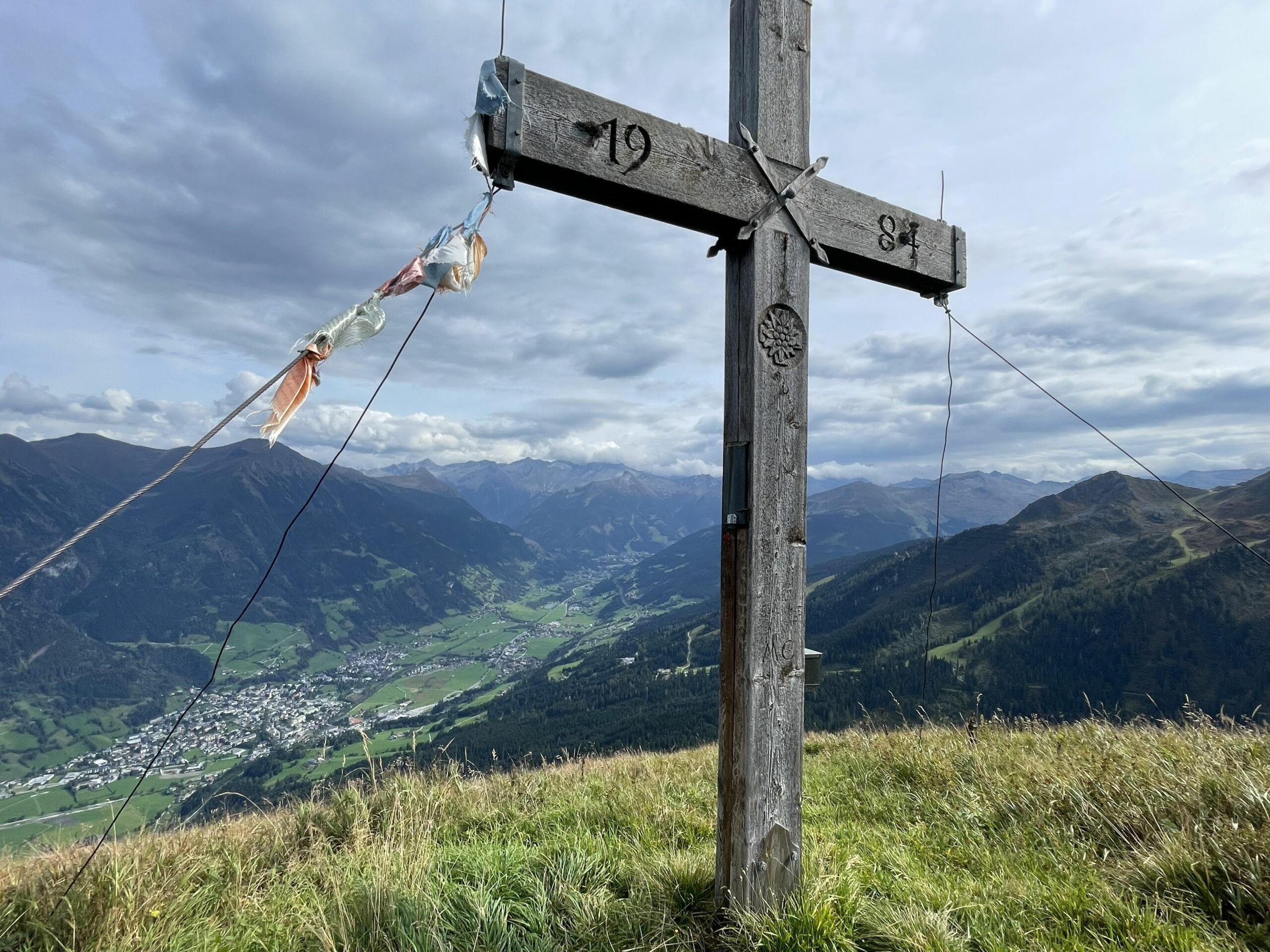 Holzgipfelkreuz auf grasigem Berg mit weitem Blick über Tal, Ortschaft und umliegende Berge unter wolkigem Himmel. – Schmaranzhütte