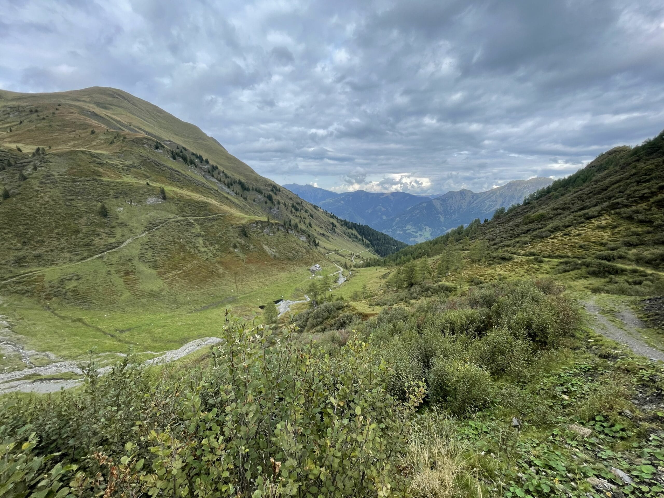 Weites alpines Tal mit schmalem Pfad, kleinem Gebäude, Bachlauf und bewaldeten Hängen unter dichter Wolkendecke.