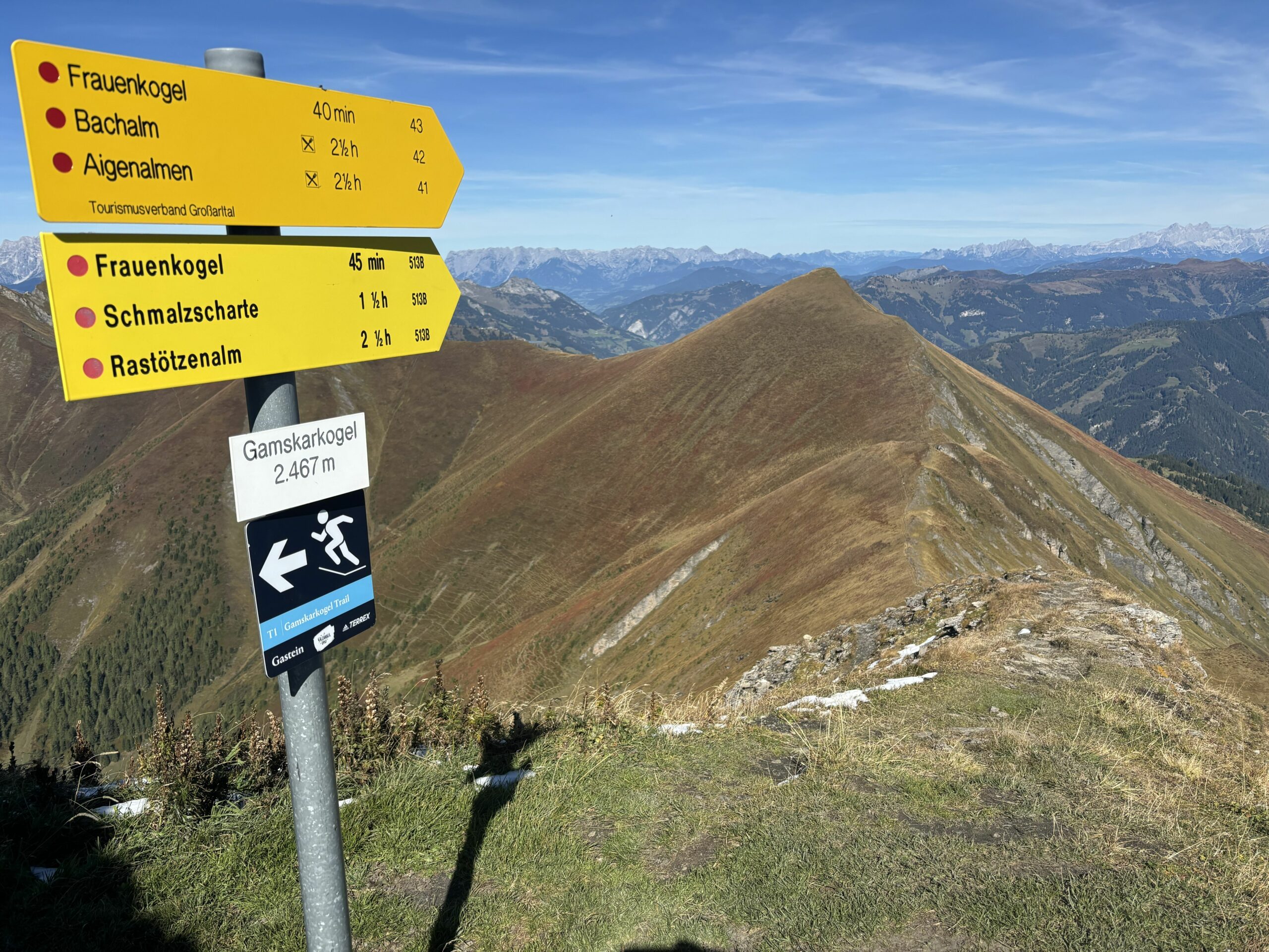 Wegweiser am Gamskarkogel auf 2.467 m vor langem Bergkamm und weiter Alpenkulisse unter blauem Himmel.