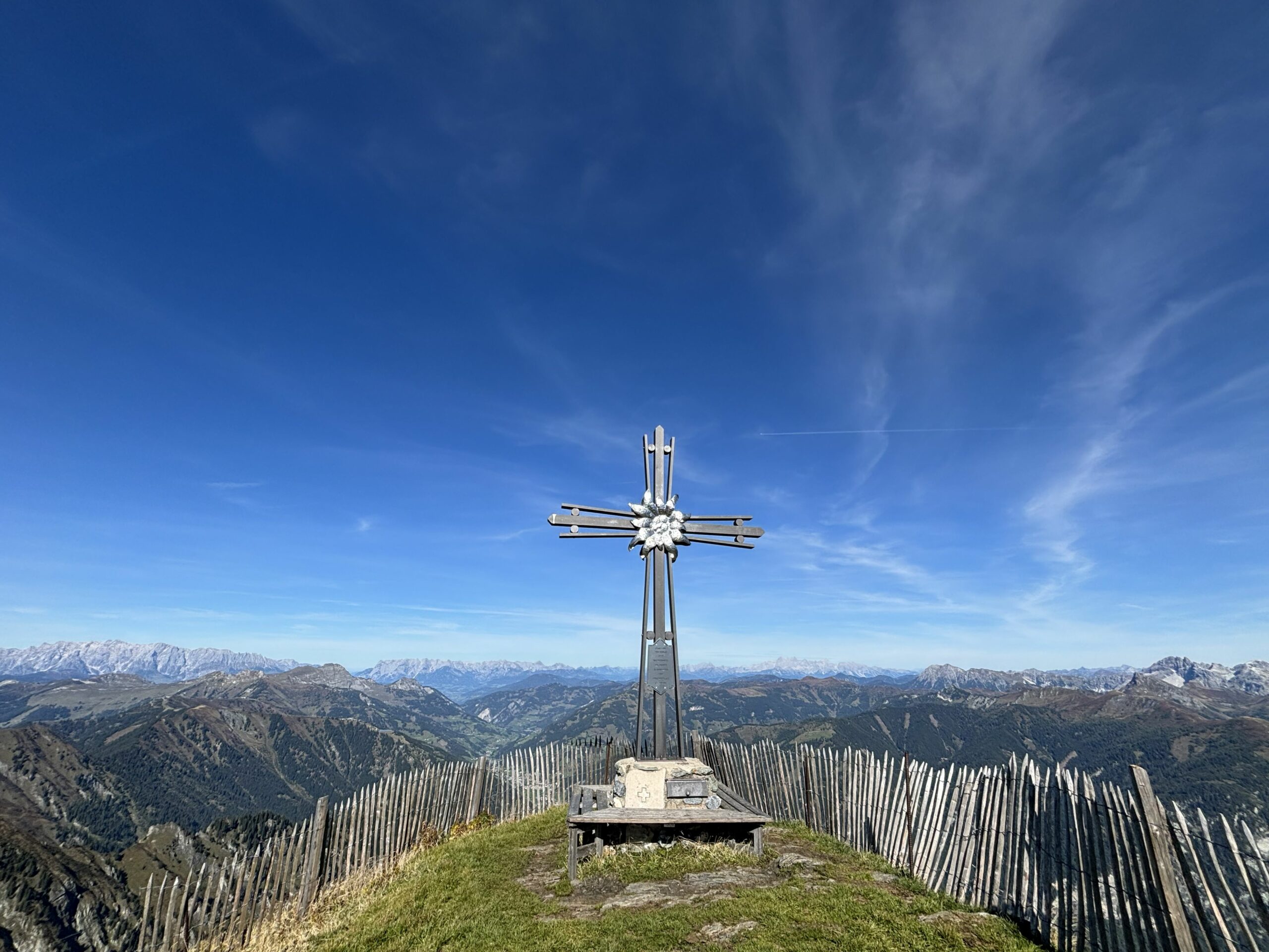Gipfelkreuz auf eingezäuntem Bergpunkt mit weitem Blick über umliegende Berge unter blauem Himmel.