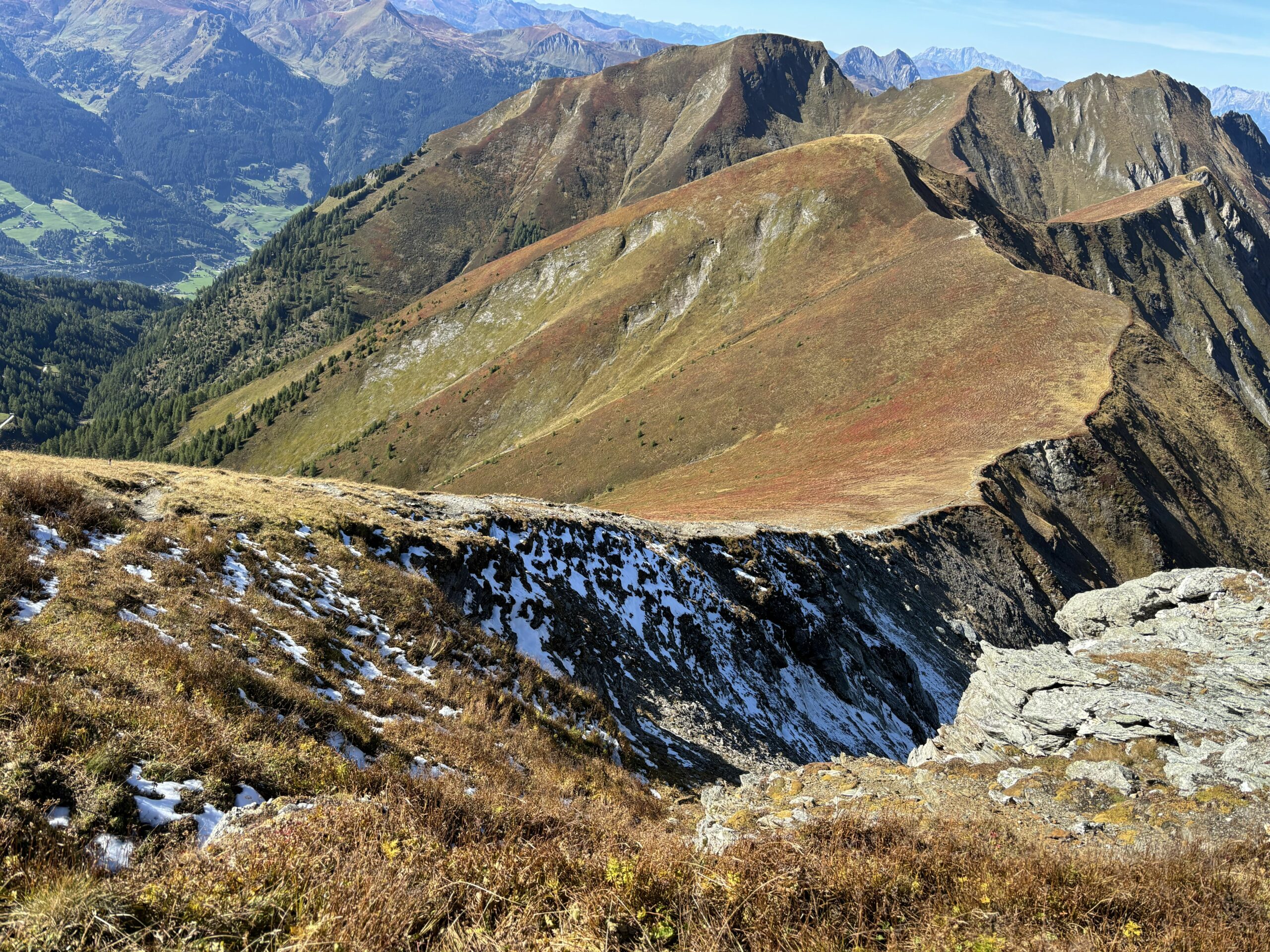 Weitblick über einen alpinen Kamm mit schmalem Pfad, steilen Hängen, etwas Altschnee und bewaldetem Tal im Hintergrund.
