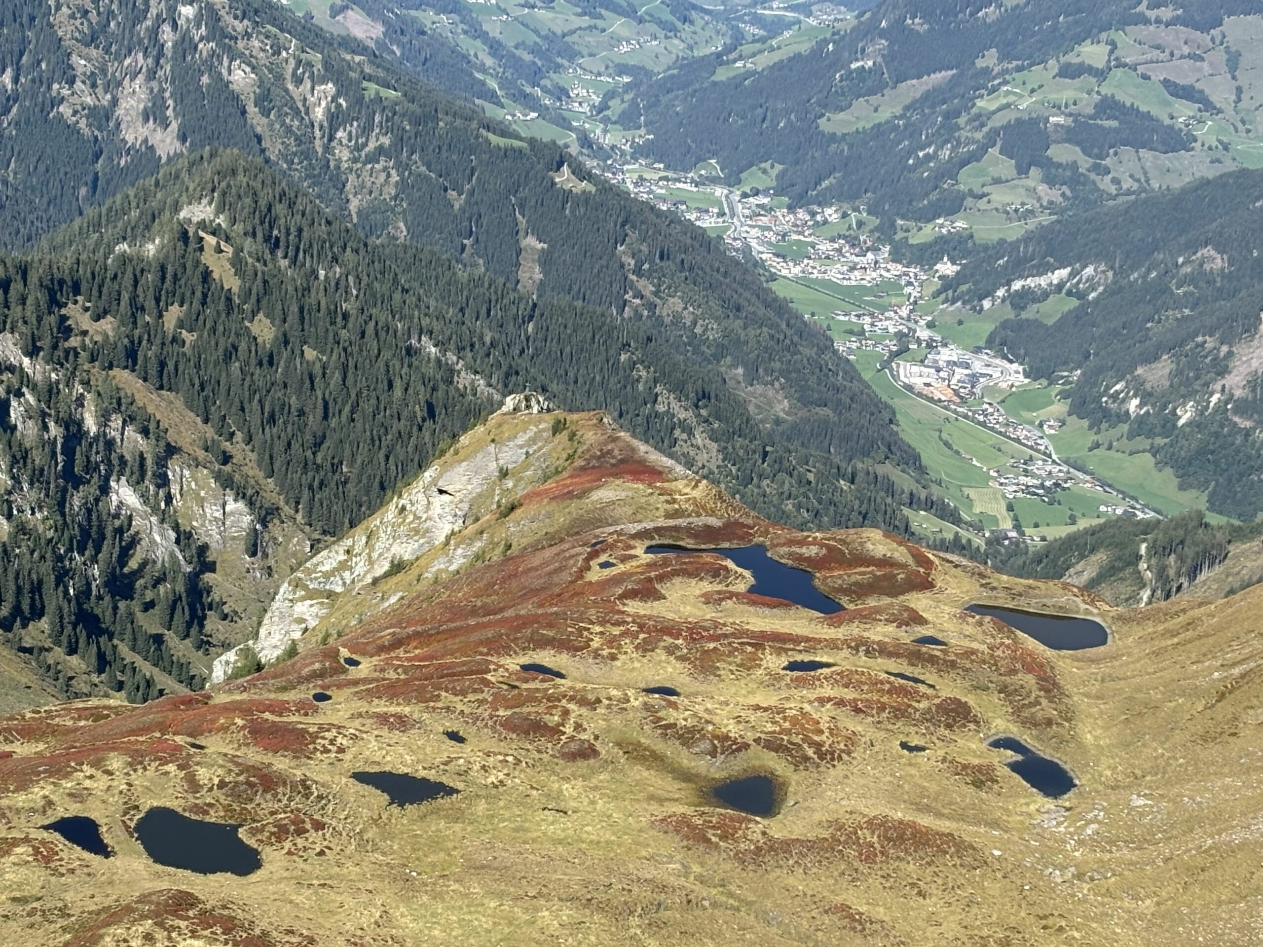 Blick von einem Höhenrücken über kleine Bergseen, bewaldete Hänge und ein Tal mit Ortschaften.