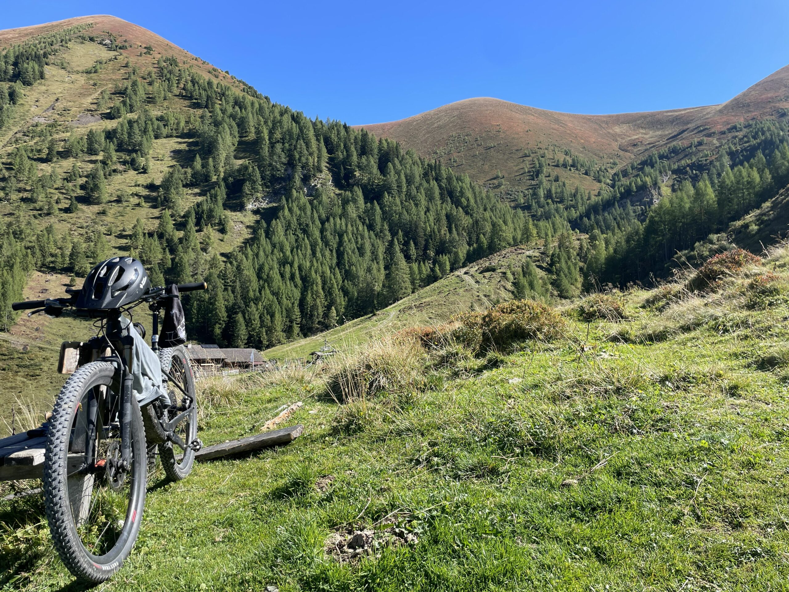 Mountainbike mit Helm auf einer Wiese vor bewaldeten Berghängen und weiten, kahlen Kuppen unter blauem Himmel.