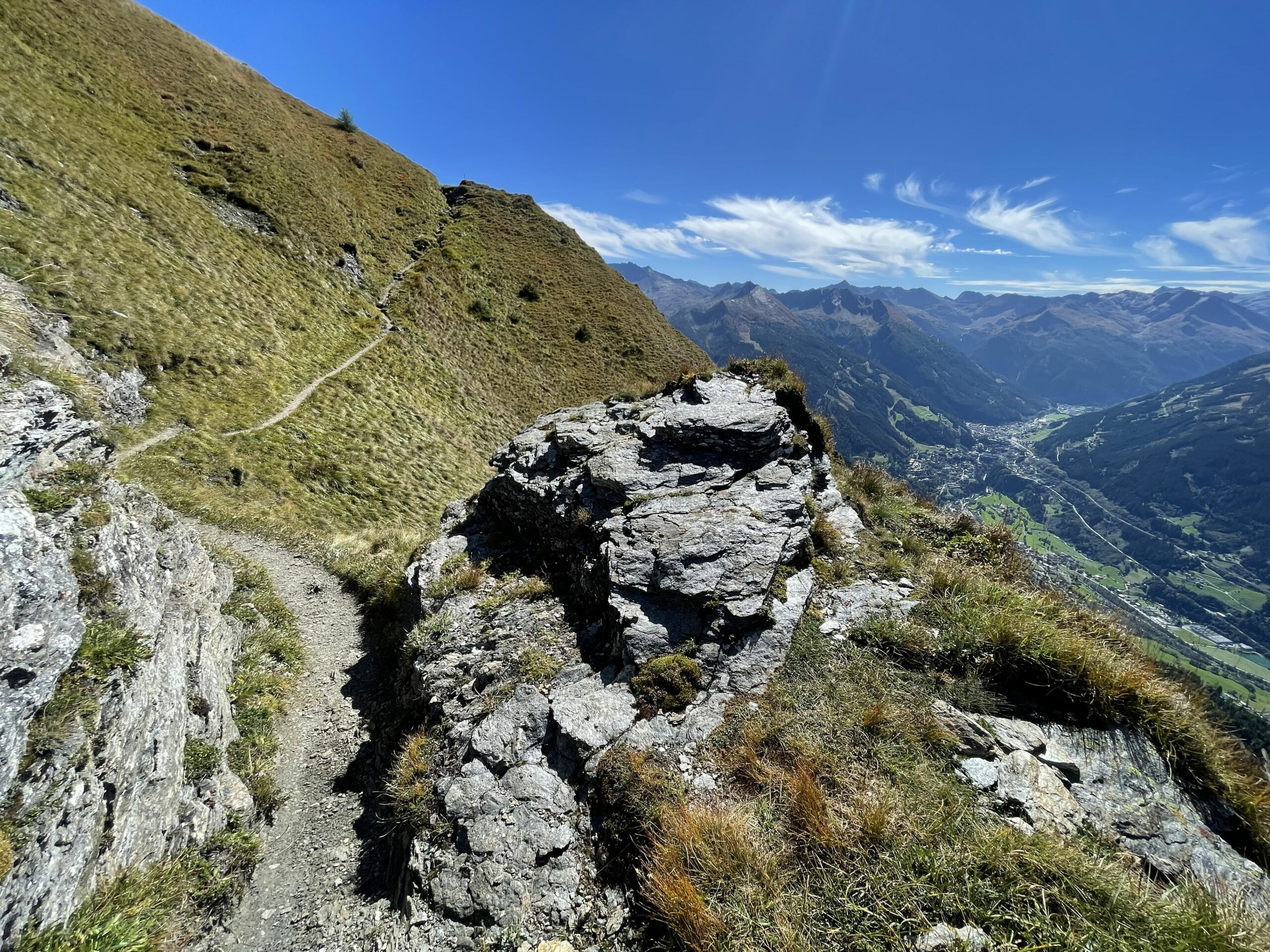 Schmaler Bergpfad quert einen grasigen Hang mit Felsen; dahinter weites Alpenpanorama über ein tiefes Tal.
