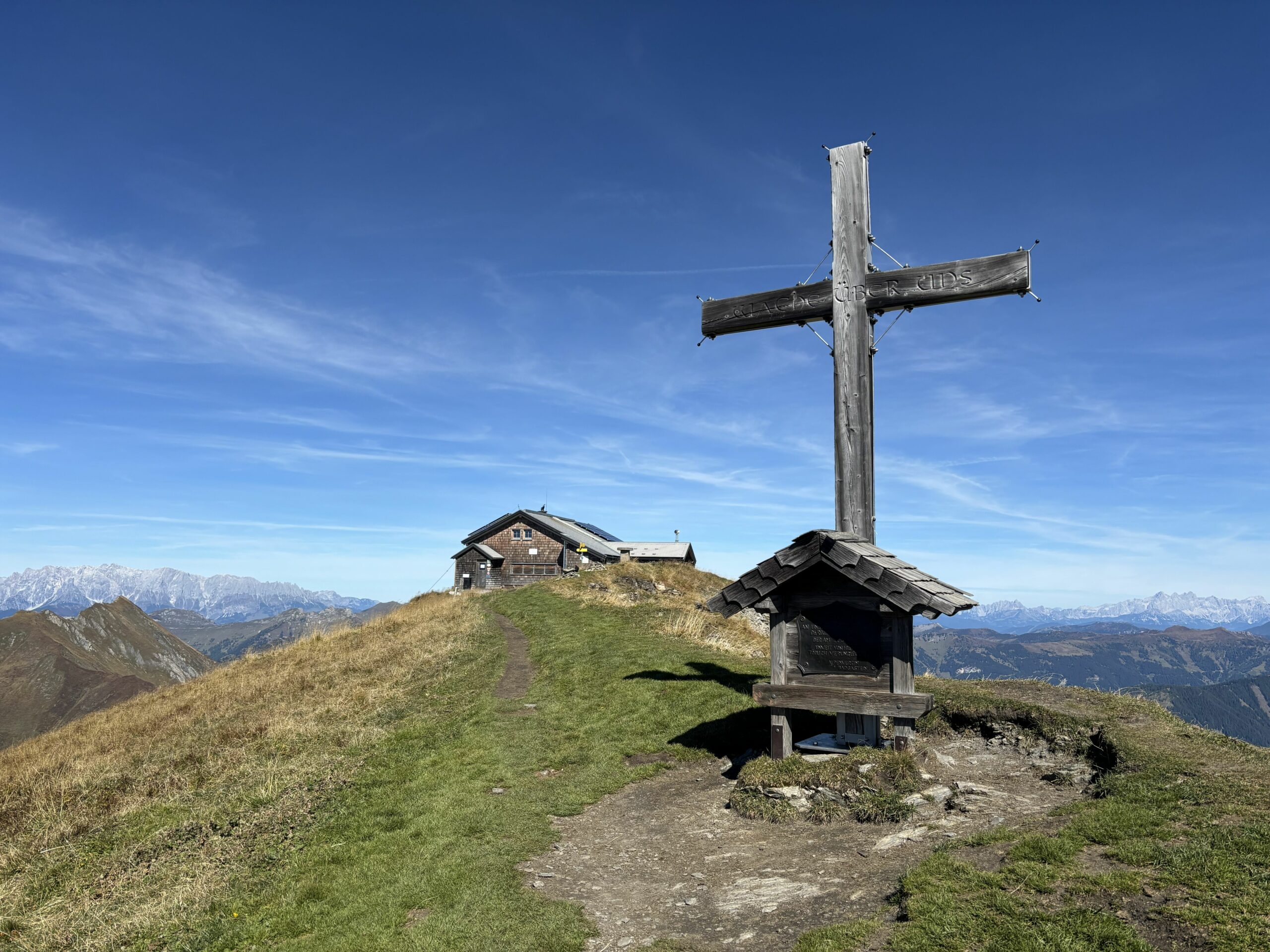 Großes Gipfelkreuz neben einer Hütte auf grasigem Bergkamm, mit schmalem Pfad und weiter Aussicht auf umliegende Berge.