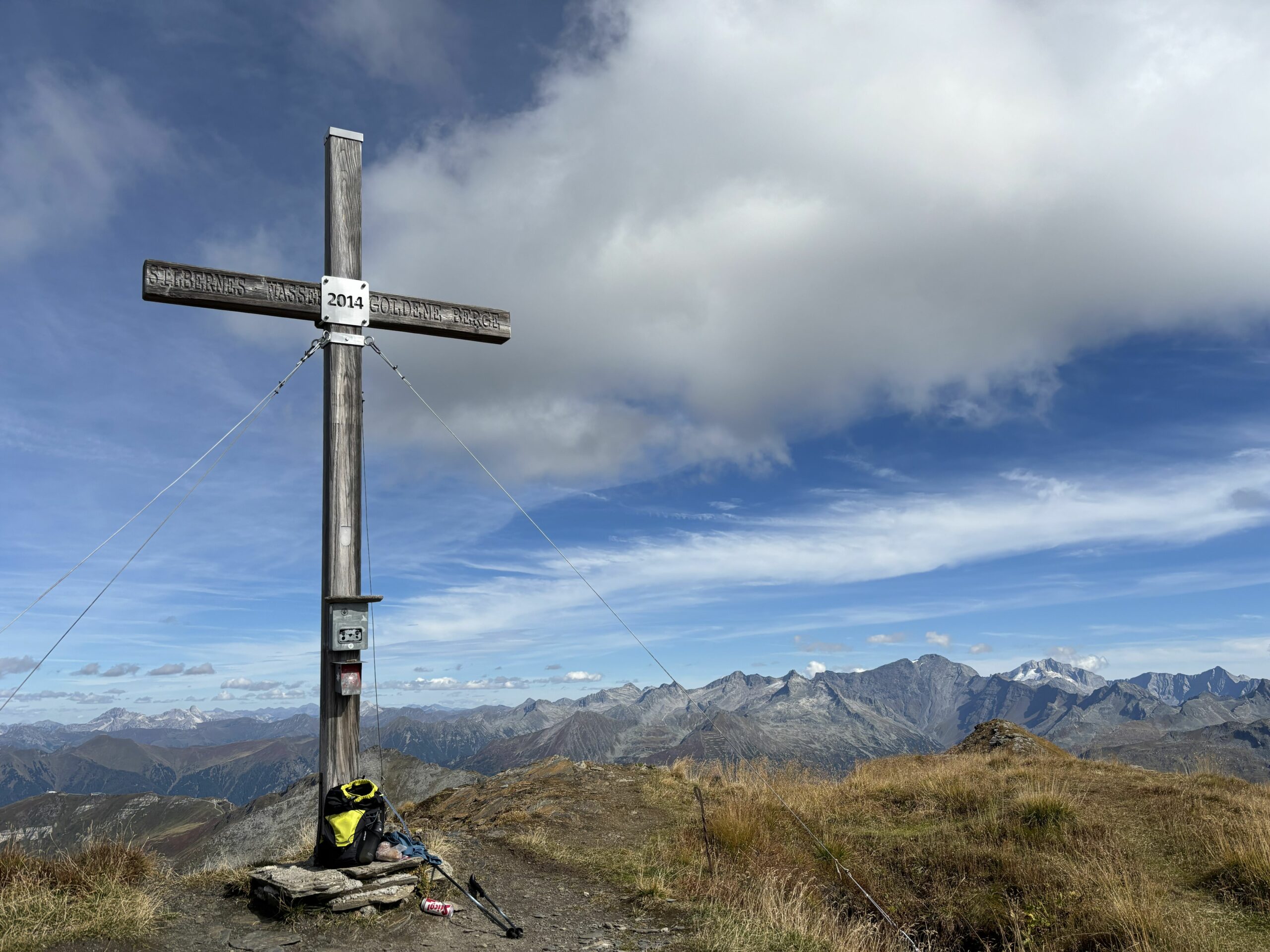 Gipfelkreuz auf einem Berg mit weitem Alpenpanorama unter wolkigem Himmel; am Fuß stehen Rucksack und Stöcke.
